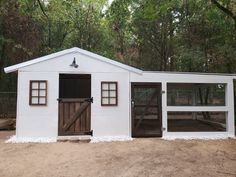 a white chicken coop with two doors and windows on the side, in front of trees