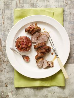 a white plate topped with meat next to a small bowl of tomato sauce on top of a green napkin