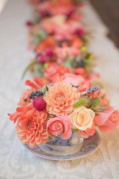 an arrangement of colorful flowers in a vase on a white table cloth lined up along the length of the table