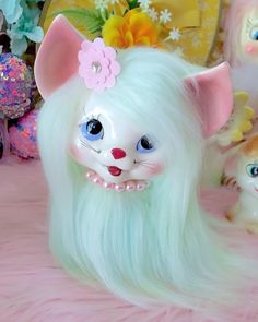 a white cat with long green hair laying on top of a pink bed next to other toys