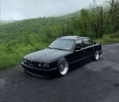 a black car parked on the side of a road near some trees and hills in the background