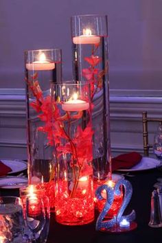 candles are lit in glass vases with red flowers and greenery on the table