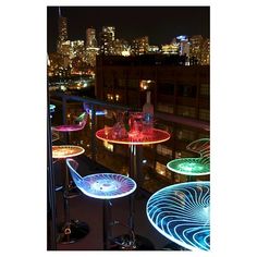 illuminated chairs and tables on a balcony overlooking the city skyline at night, lit up with colorful lights