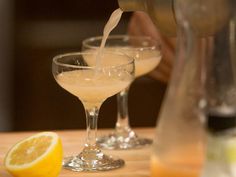 two glasses filled with liquid next to a lemon and a pitcher on a wooden table