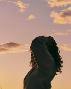 a woman standing in the sunset with her arms behind her head, looking up into the sky