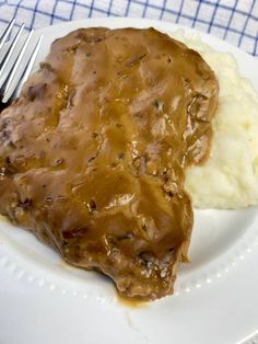 a white plate topped with meat covered in gravy and mashed potatoes next to a fork