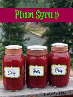 three jars of plum syrup sitting on top of a wooden table with trees in the background