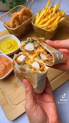 a person holding a burrito in their hand near some fries and dipping sauces