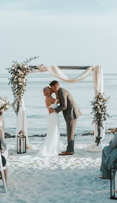 a bride and groom kissing under an arch on the beach during their wedding ceremony at sunset