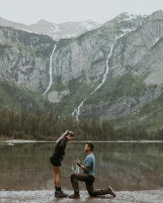 two men are sitting on the shore of a mountain lake and one is looking at his phone