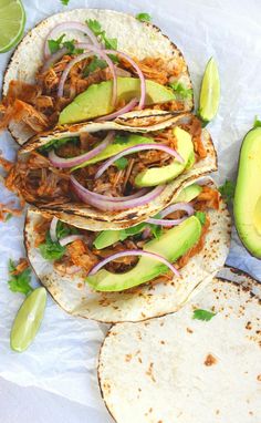 three tacos with shredded meat, onions and avocado on paper next to sliced limes