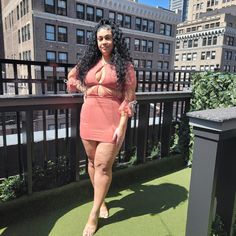 a woman standing on top of a balcony next to tall buildings in the background, wearing a pink dress and gold sandals