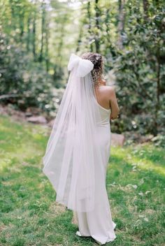 a woman in a white dress and veil standing in the grass with trees behind her