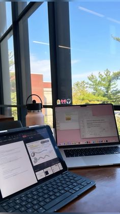 two laptops are sitting on a wooden table in front of large windows, one is open and the other is closed