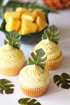 three cupcakes with white frosting and green leaves on them, next to pineapples