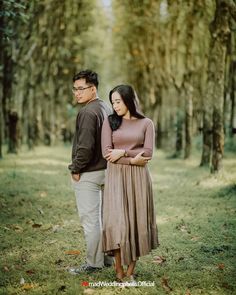 a man and woman standing next to each other in the grass with trees behind them