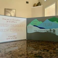 two wedding cards sitting on top of a counter next to each other, with mountains in the background