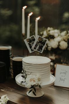 a white cake sitting on top of a wooden table next to candles and pictures with flowers