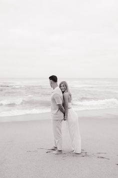 a man and woman standing on top of a sandy beach next to the ocean in black and white