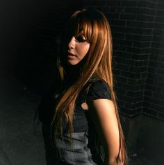 a woman with long red hair standing in front of a brick wall and looking at the camera