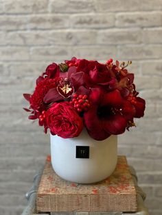 a white vase filled with red flowers sitting on top of a wooden table next to a brick wall