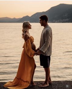 a man and woman standing next to each other near the water at sunset with mountains in the background