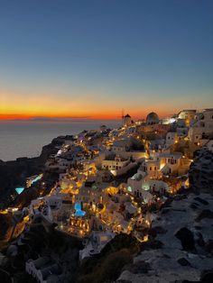 the sun is setting over some white buildings and lights on top of a hill by the ocean