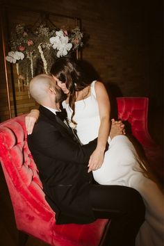 a bride and groom are sitting on a red chair in front of a flower arrangement