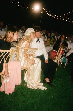 a bride and groom sitting at a dinner table in the grass with string lights overhead