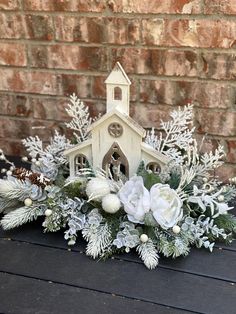 a small white church surrounded by greenery and snowflakes on a table next to a brick wall