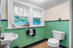 a bathroom with green and white tiles on the walls, toilet, sink and window