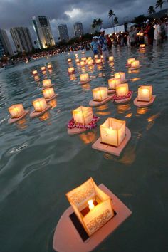 candles floating in the water at night with people standing around them and buildings in the background