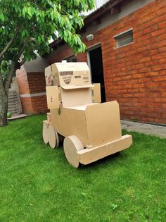 a cardboard cart sitting in the grass near a brick building with a tree on it