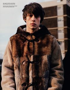 a young man wearing a fur coat in front of a building and looking at the camera