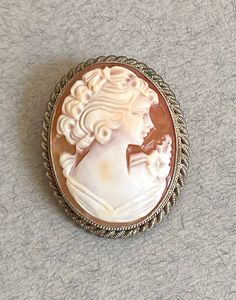 1960s rope twist edge cameo brooch featuring woman with flower corsage.  Full UK sterling silver hallmarks for the year 1966.  Signed by maker.  Excellent vintage condition.  Shell is in good order with no cracks or chips.  Clasp works well.  Gently polished, may retain some silver patina.  Weight: 7g  Presented in organza gift pouch.  Please note that any boxes you see in photos are usually just for display purposes.  To ensure there's no confusion, make sure to read the description above to se Anniversary Oval Brooches, Classic Oval Brooches, Victorian Style White Cameo Brooches, Vintage White Cameo Brooches, Luxury Vintage Cameo Brooches, White Oval Cameo Brooches, Antique Oval Cameo Brooch, Oval Silver Cameo Brooch, Silver Hallmarks