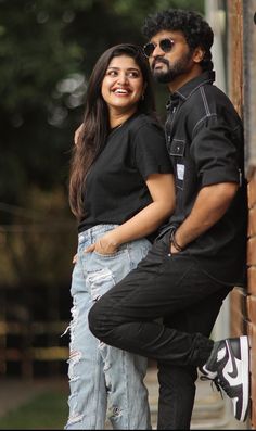 a man and woman standing next to each other near a brick wall with trees in the background