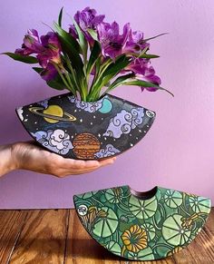 a hand holding a vase with purple flowers in it and an object shaped like a bird