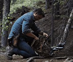 a man kneeling down next to a dead animal in the woods with trees around him