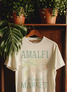 a t - shirt that says amalfi flower market hanging on a rack with potted plants