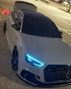 the front end of a white car parked in a parking lot at night with its lights on