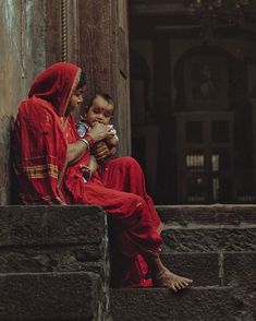 a woman and child sitting on the steps in front of an old building, one is holding her baby