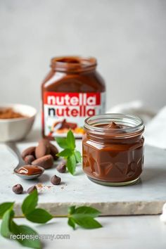 two jars of nutella and some nuts on a white surface with leaves around them