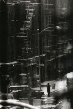 a black and white photo of traffic on a city street in the snow with fire escapes