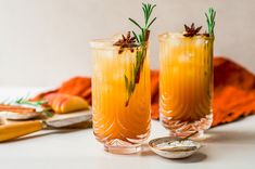 two tall glasses filled with yellow liquid and topped with rosemary garnish, sitting on a white surface