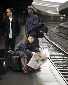 three people waiting on the train tracks with their suitcases and newspapers in front of them