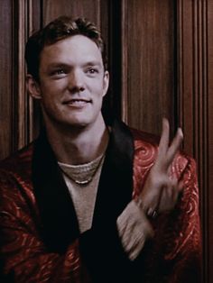 a man in a red jacket making the v sign with his hand while standing next to a wood paneled wall