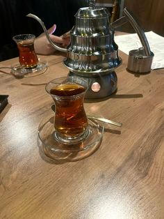 a wooden table topped with two cups of tea and a metal kettle filled with liquid