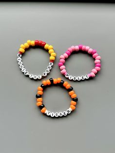 three bracelets with letters on them sitting on top of a gray surface and one has an orange, white, and black beaded name charm