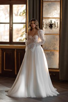 a woman in a white wedding dress standing by a window with her hands on her hips