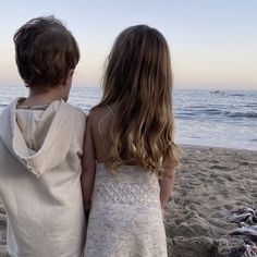 two children standing on the beach looking out at the water and sand with their backs to the camera